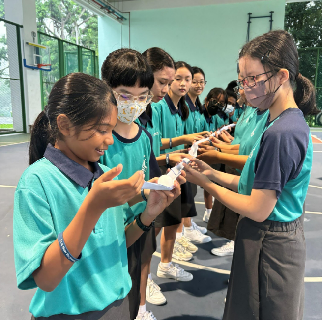 A row of students in teal uniforms pass a small paper object along a line outdoors.